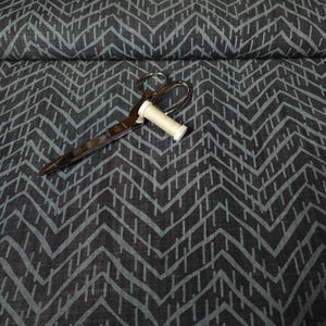 Puede incluir: Unas tijeras plateadas y una bobina de hilo blanco descansan sobre una tela gris oscuro con un patr&oacute;n de chevr&oacute;n turquesa. La tela est&aacute; doblada, lo que sugiere que es material para coser o crear.