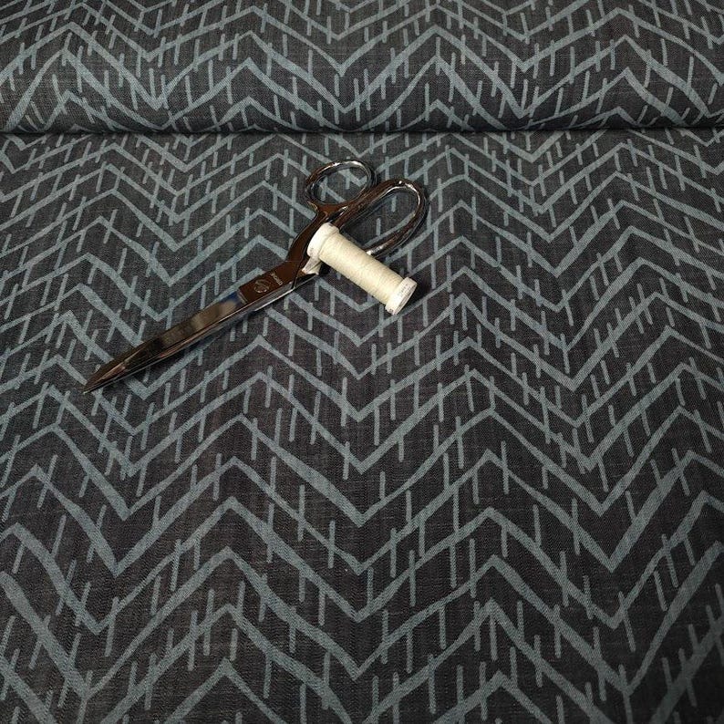 Puede incluir: Unas tijeras plateadas y una bobina de hilo blanco descansan sobre una tela gris oscuro con un patr&oacute;n de chevr&oacute;n turquesa. La tela est&aacute; doblada, lo que sugiere que es material para coser o crear.