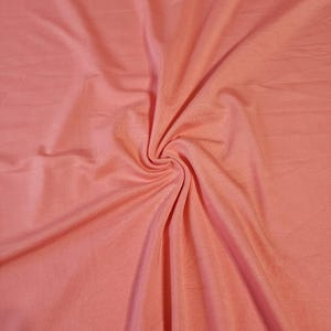 May include: A close-up shot of a peach-colored fabric with a soft, flowing texture. The fabric is gathered in a spiral, creating a pattern of folds and shadows. The material appears to be lightweight and suitable for clothing or other textile projects.