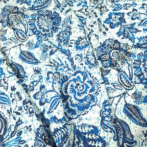 May include: Close-up of a fabric with a repeating floral pattern in shades of blue and white. The design features large, stylized flowers, leaves, and other botanical elements. The fabric appears to be cotton or a similar material, with a slightly textured surface.