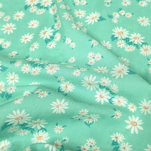 May include: A light mint green fabric featuring a repeating pattern of small white daisy flowers with yellow centers and small green leaves. The textile has a soft, draped appearance with gentle folds creating shadows across the surface.