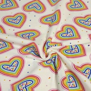 K&ouml;nnte beinhalten: Cremefarbener Stoff mit einem sich wiederholenden Muster aus regenbogenfarbenen Herzen. Jedes Herz ist rosa umrandet und mit Schichten aus Gelb, Orange, Gr&uuml;n, Blau und Lila gef&uuml;llt. Kleine, bunte Punkte sind &uuml;ber den Stoff verstreut.