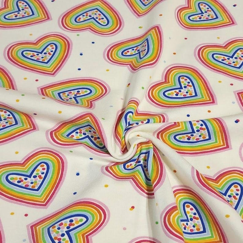 K&ouml;nnte beinhalten: Cremefarbener Stoff mit einem sich wiederholenden Muster aus regenbogenfarbenen Herzen. Jedes Herz ist rosa umrandet und mit Schichten aus Gelb, Orange, Gr&uuml;n, Blau und Lila gef&uuml;llt. Kleine, bunte Punkte sind &uuml;ber den Stoff verstreut.