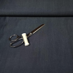 May include: A solid dark charcoal gray fabric with a subtle woven texture. Resting on the fabric is a pair of silver metal sewing scissors with a small white spool of thread positioned between the blades near the handle of the scissors.