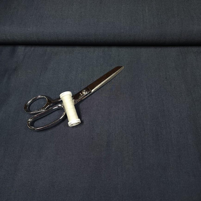 May include: A solid dark charcoal gray fabric with a subtle woven texture. Resting on the fabric is a pair of silver metal sewing scissors with a small white spool of thread positioned between the blades near the handle of the scissors.