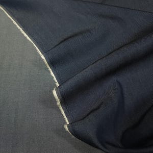 May include: A piece of dark navy blue fabric with a visible weave texture. The material is folded over to reveal a lighter, off-white fuzzy or fleece-like backing along the edge, showing a clear contrast between the smooth dark top and the soft underside.