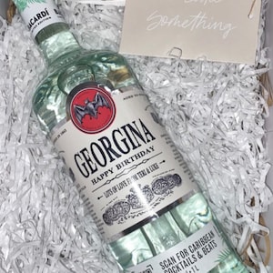 May include: A clear glass bottle of Georgina rum with a white label. The label has a bat logo and the text "Georgina Happy Birthday" with a message of love from Teri & Luke. The bottle is sitting in white shredded paper.