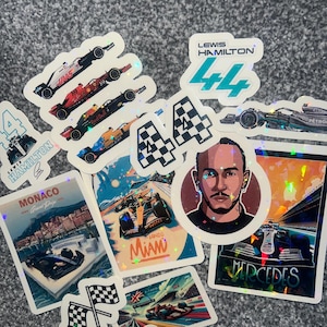 May include: A collection of holographic stickers featuring Formula 1 race cars, drivers, and race tracks. The stickers include the number 44, Lewis Hamilton, Monaco Grand Prix, and Miami Grand Prix.