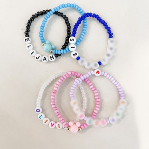 Peut inclure: Quatre bracelets en perles avec des perles et des breloques de différentes couleurs. Un bracelet porte le nom "Elijah" en perles blanches, un autre le numéro "512" en perles blanches, un autre le nom "Olivia" en perles blanches et le dernier bracelet a un charme Mickey Mouse.