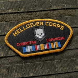 May include: A black embroidered patch with a gold border featuring the text HELLDIVER CORPS in gold, a white skull emblem, the red text CYBERSTAN CAMPAIGN, and a horizontal ribbon bar with blue, white, red, and gold stripes on a dark green fabric.