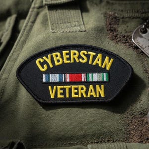 Könnte beinhalten: Ein schwarzer und gelber Aufnäher mit den Worten "CYBERSTAN VETERAN" aufgestickt. Der Aufnäher zeigt eine Reihe farbiger Streifen und ist vor einem olivgrünen Hintergrund platziert, möglicherweise eine Militäruniform.