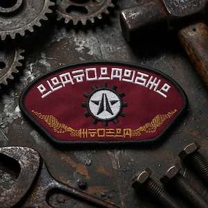 May include: A maroon embroidered patch with a black border, featuring a central white circle with a black gear and star emblem. White text arches above the emblem, while gold-colored wing-like embroidery and additional text appear below.