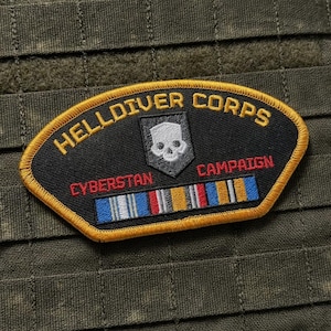 以下が含まれることがあります： 金色の縁取りが施された黒い刺繍ワッペン。金色の「HELLDIVER CORPS」と「CAMPAIGN」、赤色の「CYBERSTAN」の文字、白いドクロのエンブレム、青、白、赤、黄、灰色のストライプが入ったマルチカラーのリボンバーがデザインされています。
