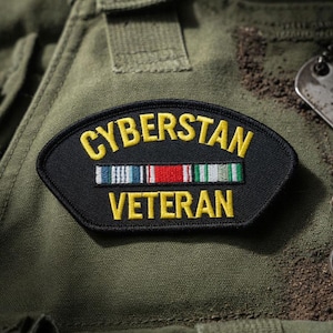 Może przedstawiać: Czarna i żółta naszywka z wyhaftowanymi słowami "CYBERSTAN VETERAN". Naszywka posiada szereg kolorowych pasków i jest umieszczona na oliwkowym tle, prawdopodobnie na mundurze wojskowym.