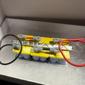 May include: A rectangular arrangement of yellow battery cells with silver connectors and grey plastic clips. Black and red wires are connected to the assembly, which is inside a white box.
