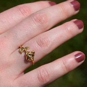 Pu&ograve; includere: Un anello dorato con un design a forma di rosa. L'anello &egrave; indossato su un dito, con il dettaglio della rosa in cima. Lo sfondo &egrave; un verde sfocato, che suggerisce un ambiente esterno. Il design delicato dell'anello &egrave; il punto focale.