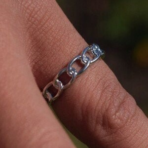 Può includere: Un anello a catena d'argento indossato su un dito. L'anello è composto da piccoli anelli interconnessi, creando un aspetto a catena. L'anello è un gioiello semplice ed elegante.