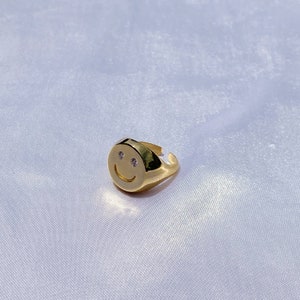 Può includere: Un anello color oro con un design a faccina sorridente. L'anello presenta un viso rotondo con due piccole pietre trasparenti per gli occhi e una linea curva per la bocca. L'anello è posto su uno sfondo bianco.
