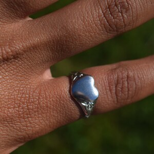 Può includere: Un anello d'argento con un design a forma di cuore. L'anello è indossato su un dito, con dettagli decorativi attorno al cuore. Lo sfondo è verde sfocato, suggerendo un ambiente esterno. Un gioiello.