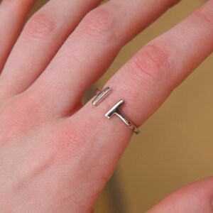 Può includere: Un anello aperto color argento dal design minimalista. L'anello presenta due elementi diritti a forma di barra che non si collegano completamente, creando un design aperto. L'anello è indossato su un dito.