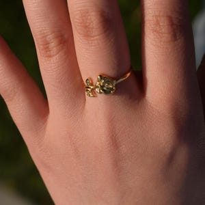 Pu&ograve; includere: Un anello dorato con un motivo floreale, indossato su un dito. L'anello presenta un design di fiori e foglie. La mano &egrave; a fuoco, con uno sfondo sfocato di tonalit&agrave; verdi e blu. Un gioiello delicato.