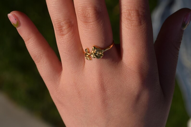 Pu&ograve; includere: Un anello dorato con un motivo floreale, indossato su un dito. L'anello presenta un design di fiori e foglie. La mano &egrave; a fuoco, con uno sfondo sfocato di tonalit&agrave; verdi e blu. Un gioiello delicato.