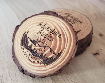 May include: Two wooden coasters with a natural bark edge. Each coaster is engraved with the words "Adventure Awaits" above a mountain and sun design. The coasters are made from a cross-section of a tree branch.