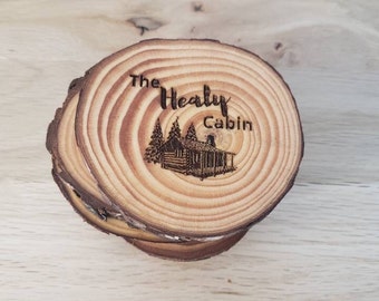May include: Set of four wooden coasters with a rustic design. The coasters feature a cabin with trees and the text "The Healy Cabin".