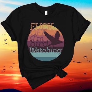 May include: Black t-shirt with a circular graphic featuring a bird silhouette and the text "FUCK OFF I'm bird Watching." The design includes a sunset background with birds in flight, creating a nature-themed design.