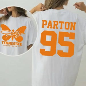 May include: A white t-shirt featuring orange graphics. The back displays the text PARTON and the number 95 in a large varsity font. The front, shown in a circular inset, features an orange butterfly graphic with the text Dolly, TENNESSEE, FOOTBALL.