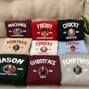 May include: A collection of nine folded sweatshirts in various colors, each featuring a horror movie character graphic, the character's name in a distressed white font, an establishment year, and the corresponding movie title printed below.