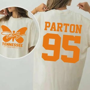 May include: An off-white t-shirt featuring orange graphics. The back displays the text PARTON above the number 95 in large block letters. The front shows an orange butterfly graphic with the text Dolly, TENNESSEE, and FOOTBALL.
