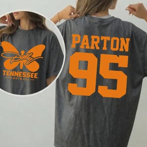 May include: A dark gray t-shirt featuring orange graphics. The back displays the text PARTON above the number 95 in a bold font. The front circular inset shows an orange butterfly graphic with the text Dolly Tennessee Football.