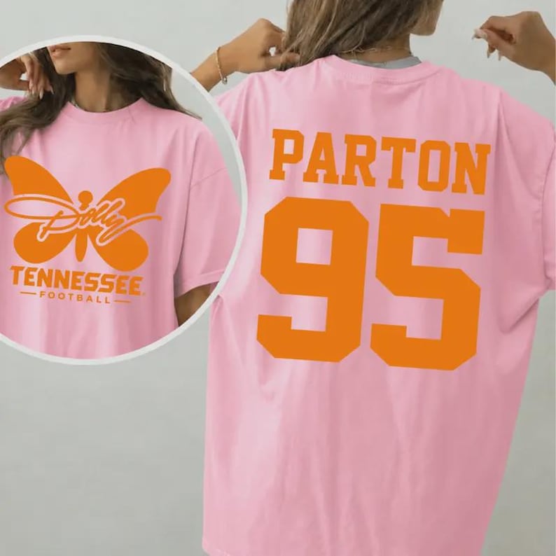 May include: A light pink t-shirt featuring a large orange butterfly graphic on the front with the text Dolly Tennessee Football. The back of the shirt displays the name Parton in large orange letters above the large orange jersey number 95.