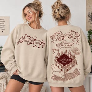 May include: A beige crewneck sweatshirt featuring dark red graphic prints. The front displays a banner with the text 'I solemnly swear that I am up to no good' and footprints. The back shows a detailed illustration of a castle and map.