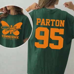 May include: A dark green t-shirt featuring an orange graphic on the front with a butterfly, the text Dolly, Tennessee, and Football. The back of the t-shirt displays the orange text Parton and the large orange number 95 in a block athletic font.