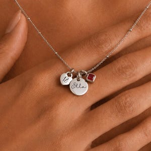 Peut inclure: Un collier argenté doté d'une chaîne délicate avec de petites perles. Trois breloques pendent au centre : un petit cœur gravé du nombre 18, un disque circulaire gravé du prénom Ella et une petite breloque carrée avec une pierre rouge.