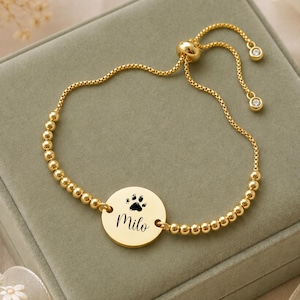 May include: A gold-toned adjustable bracelet featuring a circular charm engraved with a black paw print and the name Milo. The bracelet is constructed with small metallic beads and includes a sliding mechanism with two small clear stones at the ends.