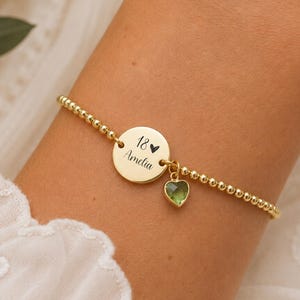 May include: A gold-toned beaded bracelet featuring a circular charm engraved with the text 18 and Amelia, accompanied by a small, faceted, heart-shaped green gemstone pendant hanging from the central disc.