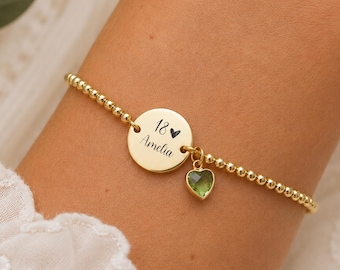 May include: A gold-toned beaded bracelet featuring a circular charm engraved with the text 18 and Amelia, accompanied by a small, faceted, heart-shaped green gemstone pendant hanging from the central disc.