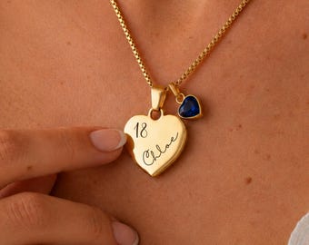 May include: A gold-toned necklace featuring a heart-shaped pendant engraved with the number 18 and the name Chloe in script. A smaller, dark blue heart-shaped charm is attached to the same bail, suspended from a delicate gold-toned box chain.