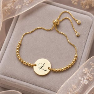 Puede incluir: Una pulsera ajustable de tono dorado con un disco circular central grabado con la letra L en letra cursiva. La pulsera está compuesta por pequeñas cuentas redondas en una cadena delicada con un cierre deslizante y dos pequeños dijes colgantes.