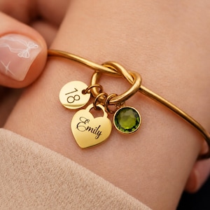 Op de afbeelding: Een goudkleurige armband met een knoopontwerp en drie bungelende bedels: een ronde schijf gegraveerd met het getal 18, een hartvormige hanger gegraveerd met de naam Emily en een ronde, gefacetteerde olijfgroene edelsteenbedel.