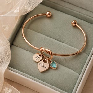 May include: A rose gold-toned metal bangle bracelet featuring a knot detail and three charms: a round disc engraved with the number 30, a heart-shaped charm engraved with the name Olivia, and a small, faceted light blue heart-shaped crystal charm.