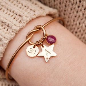 Può includere: Bracciale rigido color oro con un design a nodo e diversi ciondoli. I ciondoli includono una gemma rossa rotonda, una stella con la lettera "A" e una piccola gemma, e un ciondolo rotondo con il numero "30".