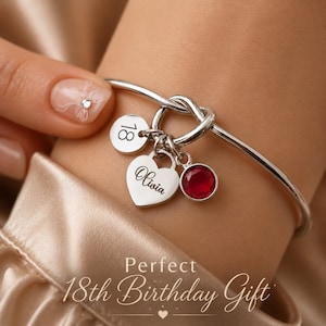 May include: A silver-toned bangle bracelet featuring a knot detail and three charms: a circular disc engraved with the number 18, a heart-shaped charm engraved with the name Olivia, and a circular charm set with a single round red gemstone.