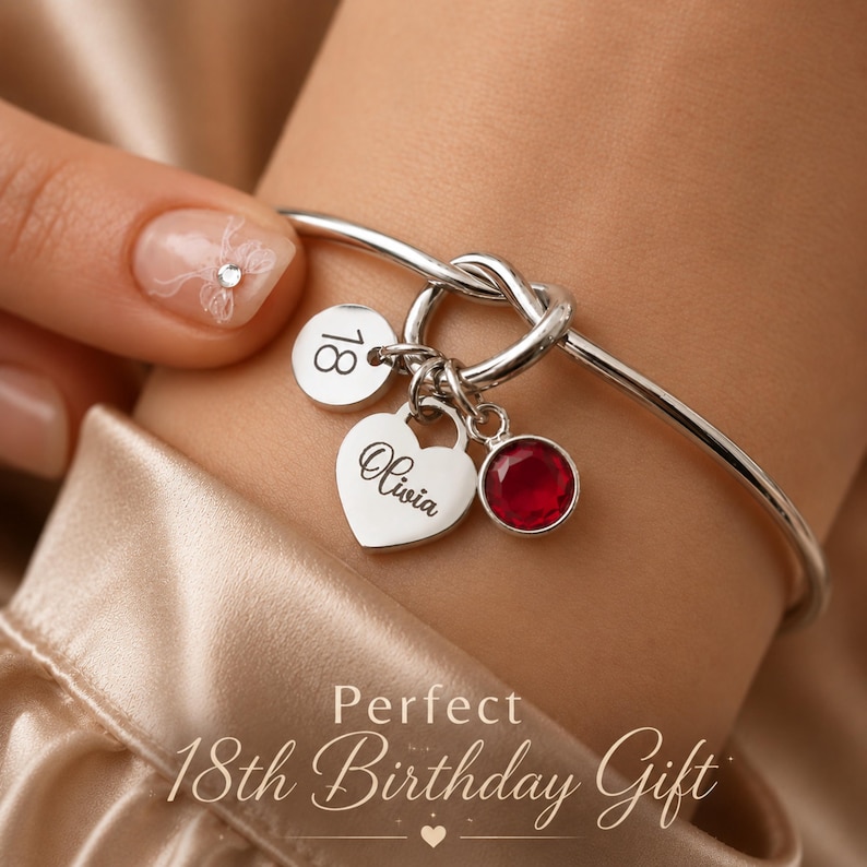 May include: A silver-toned bangle bracelet featuring a knot detail and three charms: a circular disc engraved with the number 18, a heart-shaped charm engraved with the name Olivia, and a circular charm set with a single round red gemstone.