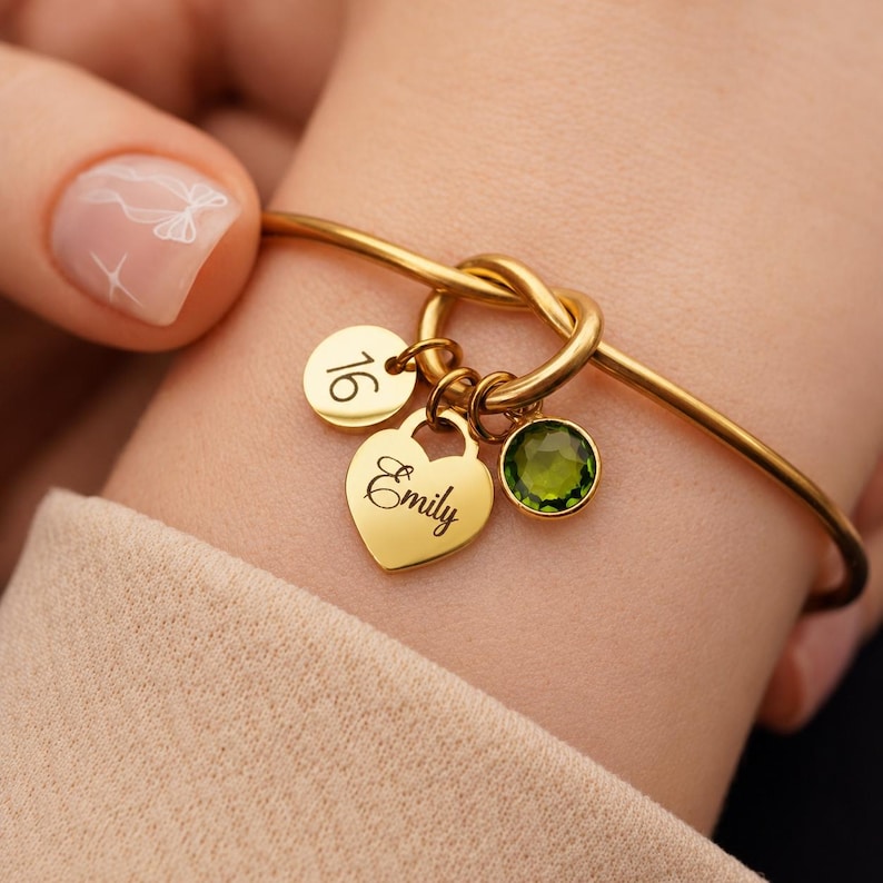 May include: A gold-toned bangle bracelet featuring a central knot design. Three charms are attached: a circular disc engraved with the number 16, a heart-shaped charm engraved with the name Emily, and a round, faceted olive-green gemstone charm.