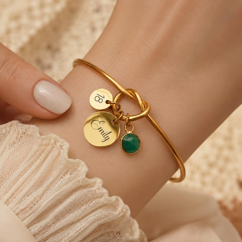 May include: A gold-toned bangle bracelet featuring a central knot design. Three charms hang from the knot: a small circular disc engraved with the number 18, a larger circular disc engraved with the name Emily, and a round faceted green gemstone.
