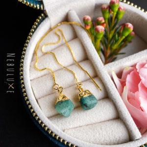 May include: A pair of gold-plated earrings with green gemstone drops. The earrings are in a jewelry box with a blue velvet lining.